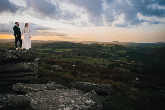 Bride and groom standing on a rocky cliff with scenic landscape during sunset, showcasing one of the best wedding photographs.