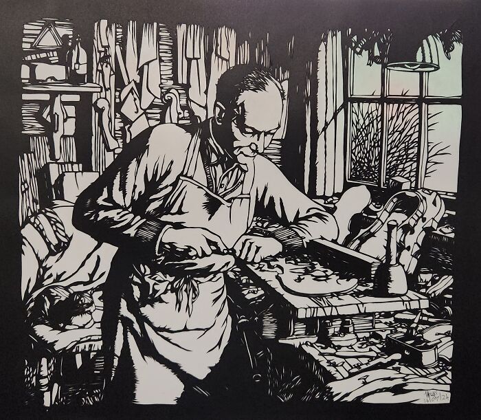 Man hand-cutting intricate paper artwork with scalpel in a busy workshop.