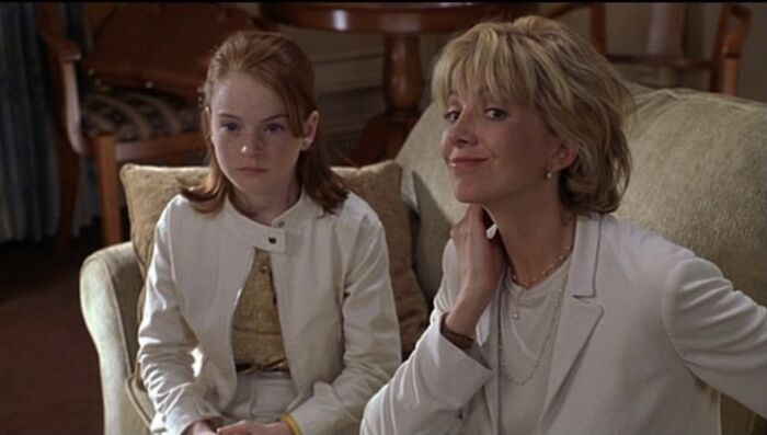 Two celebrities in the 90s sitting on a couch, one smiling and the other looking serious, both in light-colored outfits.