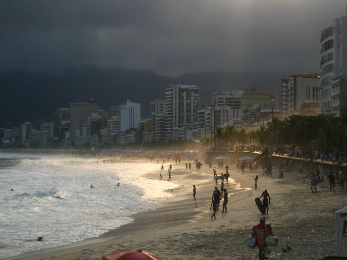 Retro Brazilian beach scene with people strolling along the shore, framed by city buildings and a moody sky.