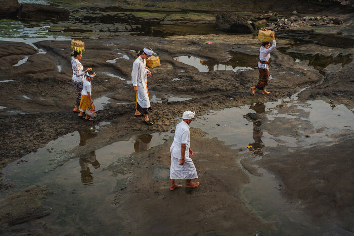 Balinese people in traditional attire celebrate Galungan by walking on a rocky path, carrying offerings.