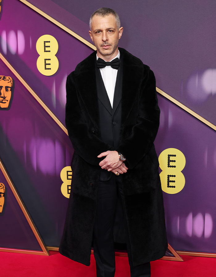 Man in a tuxedo with a fur coat on the BAFTA red carpet, showcasing daring outfits and style.