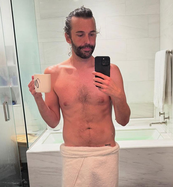 Queer Eye star in a bathroom, wrapped in a towel, holding a phone and a mug, showing weight loss transformation.