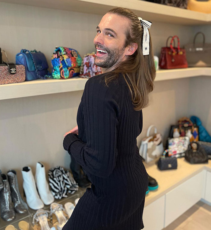 Jonathan Van Ness smiling, showcasing weight loss in a black outfit, standing in front of a shelf with colorful bags and shoes.