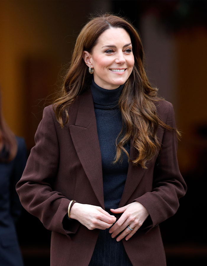 Kate Middleton in a brown coat, smiling during a public appearance related to her fashion policy.
