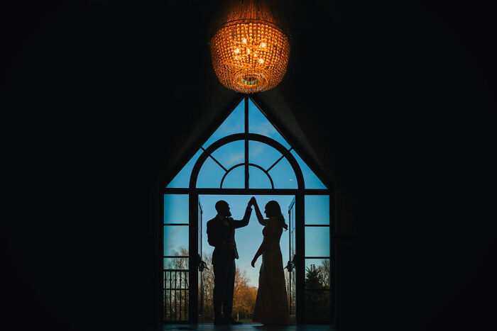 Silhouette of a couple dancing under a chandelier, framed by an arched window in a wedding photograph.
