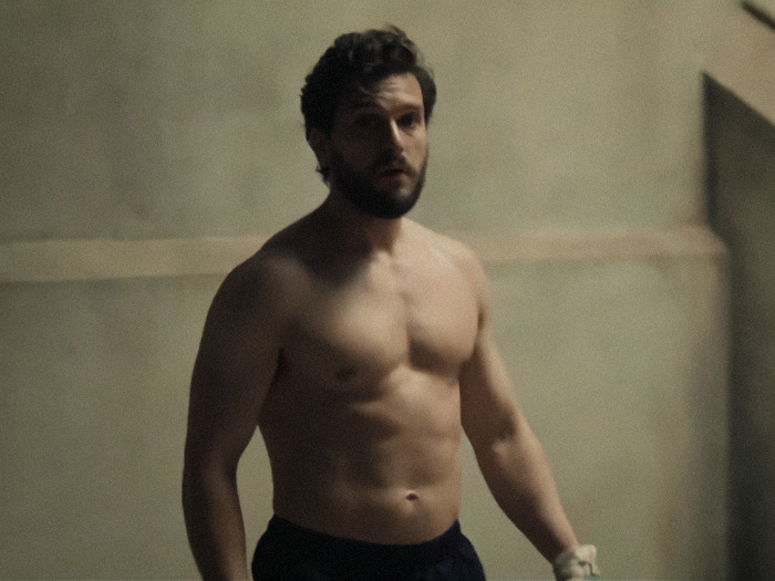 Kit Harington standing in a dimly lit room, showcasing natural physique, rejecting prosthetics on screen.