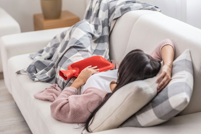 Woman relaxing on couch with hot water bottle, related to period tracker debate in mobile game.