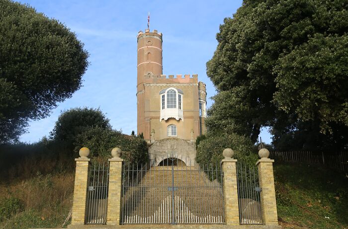 A charming, seemingly useless building with a round tower and decorative windows, surrounded by trees and a gated entrance.