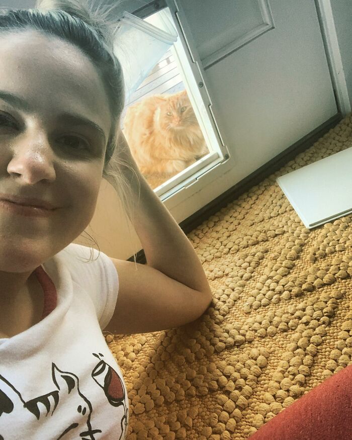 Smiling person indoors as a fluffy orange cat looks through the door flap, embracing house cat life.