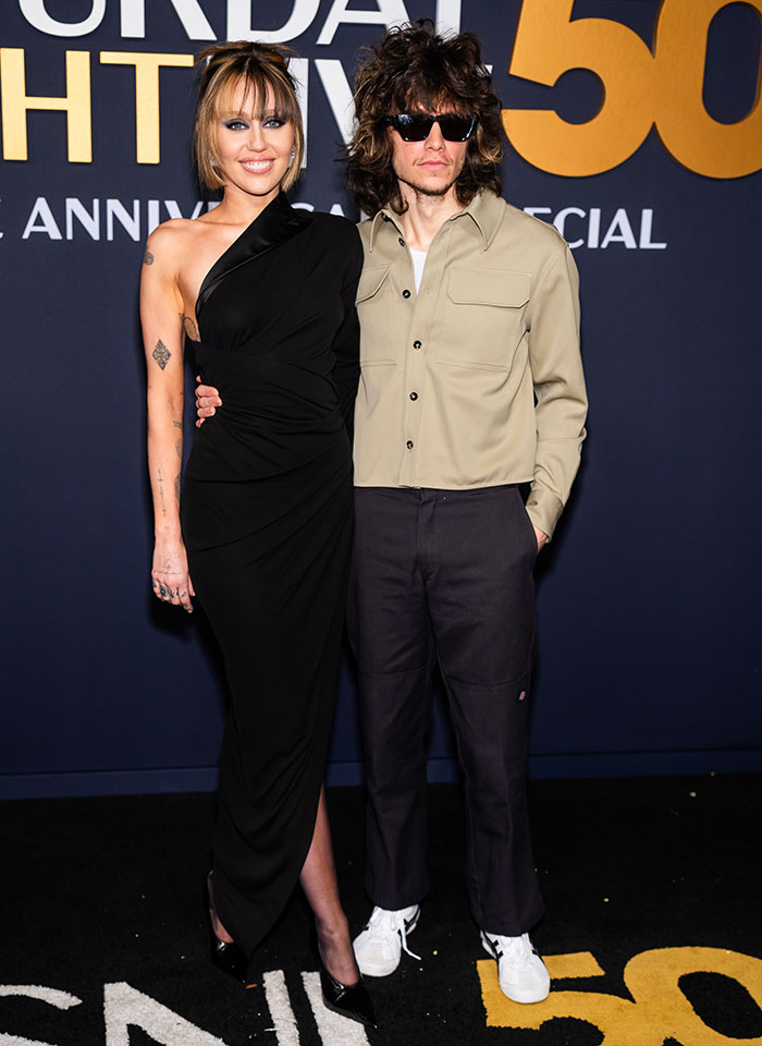 Two individuals posing on the SNL 50th anniversary red carpet in unique outfits.
