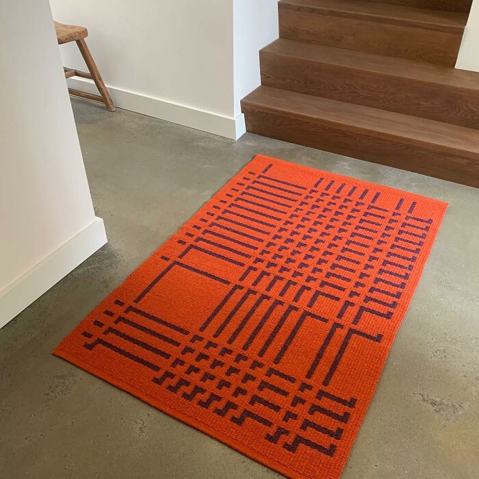 Orange geometric weave art rug creating optical illusions on a concrete floor near wooden stairs.