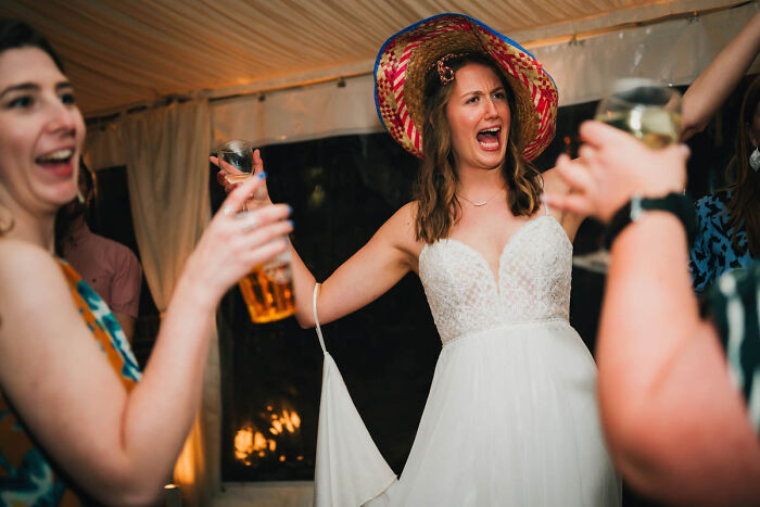 Bride joyfully wearing a colorful hat, holding a drink, celebrating at a wedding reception.