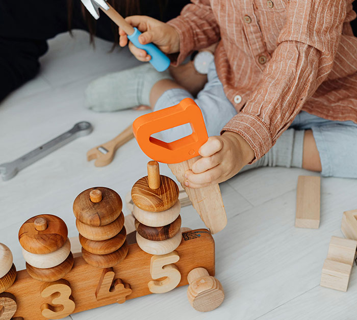 Child playing with wooden toys on the floor, holding an orange toy saw, symbolizing birthday party preparations. Child playing with wooden toys on the floor, holding an orange toy saw, symbolizing birthday party preparations.