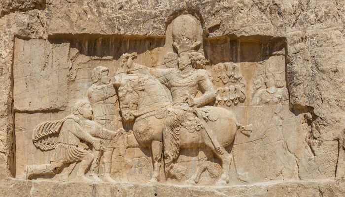 Ancient stone carving depicting a surprising war victory with figures on horseback and a soldier bowing.