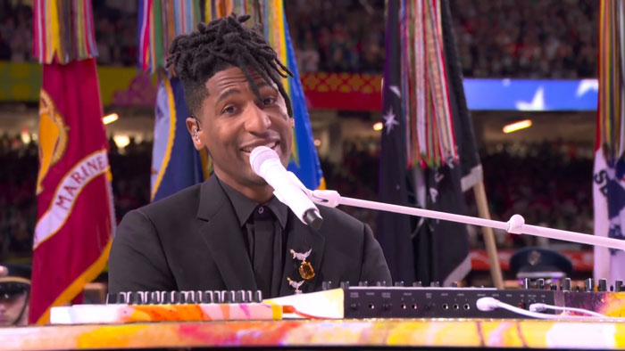 Performer singing the national anthem at Super Bowl LIX, standing before a colorful display of flags. Performer singing the national anthem at Super Bowl LIX, standing before a colorful display of flags.