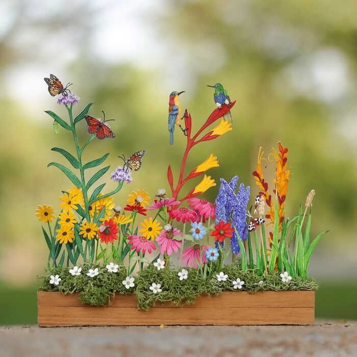 Miniature realistic paper art of animals, featuring butterflies and birds among colorful flowers on a wooden base.
