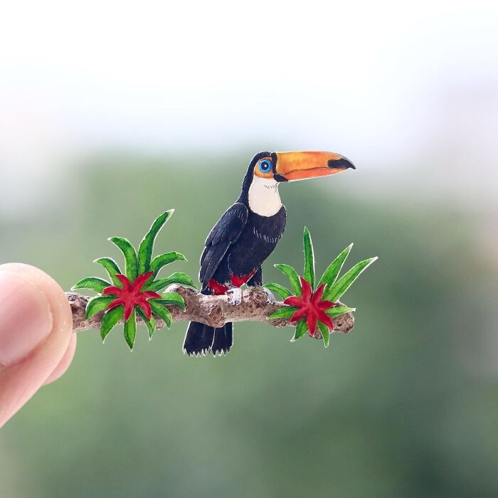 Miniature realistic paper art of a toucan perched on a branch with red flowers, created by artists to inspire conservation.