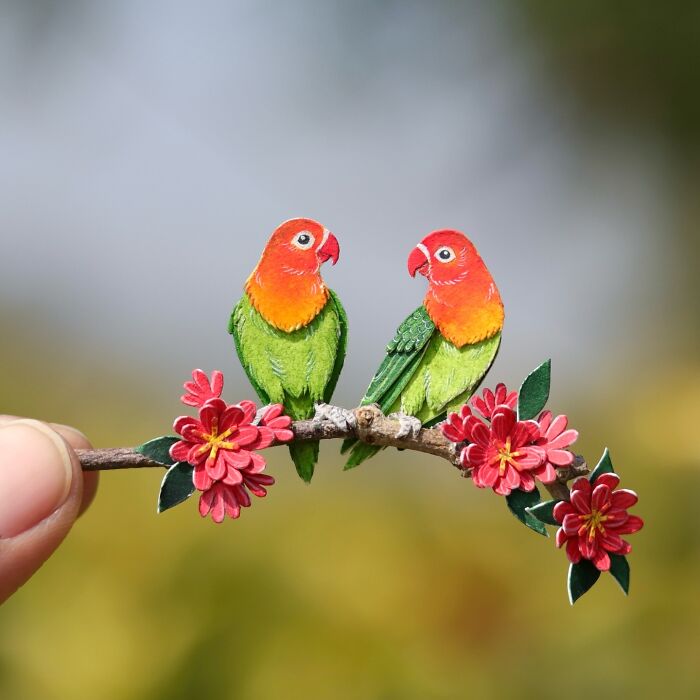Miniature realistic paper art of two colorful birds perched on a branch with red flowers.