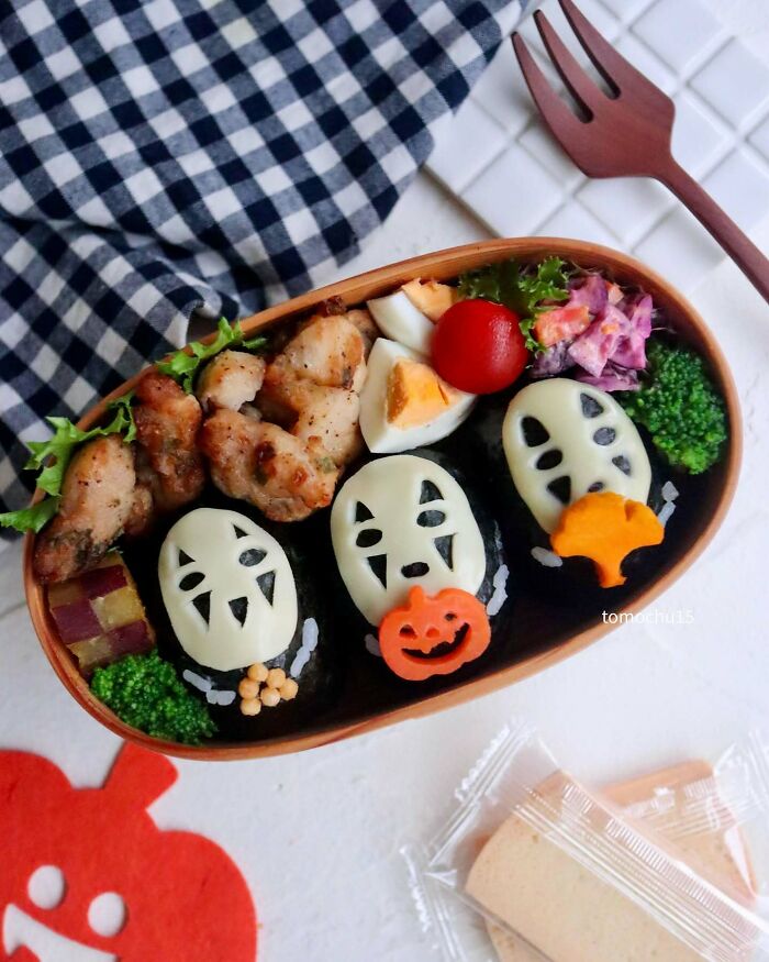 Creative lunchbox with character-themed rice balls, grilled chicken, boiled egg, tomato, broccoli, and colorful garnish.