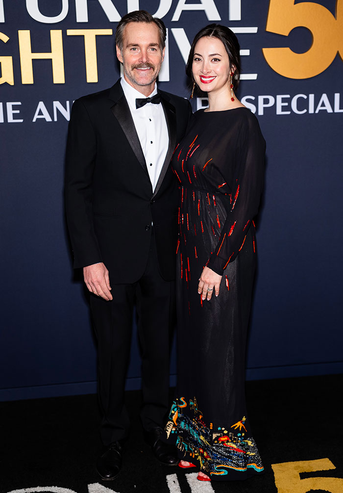 Couple in formal attire at SNL 50th anniversary red carpet, with man in tuxedo and woman in a black dress with colorful patterns.