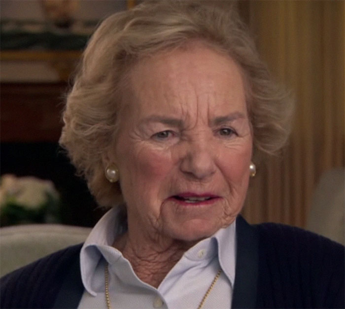 Elderly woman in a living room, wearing a pearl necklace and earrings, linked to secret Epstein documents.