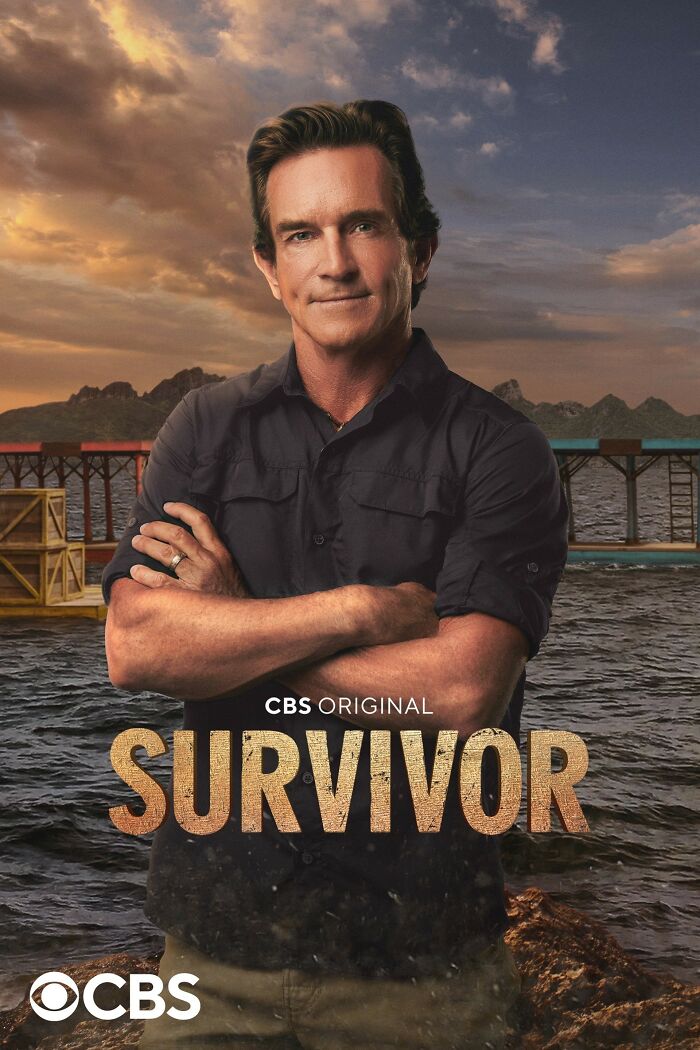 Show host stands with arms crossed in front of island backdrop for TV series "Survivor."