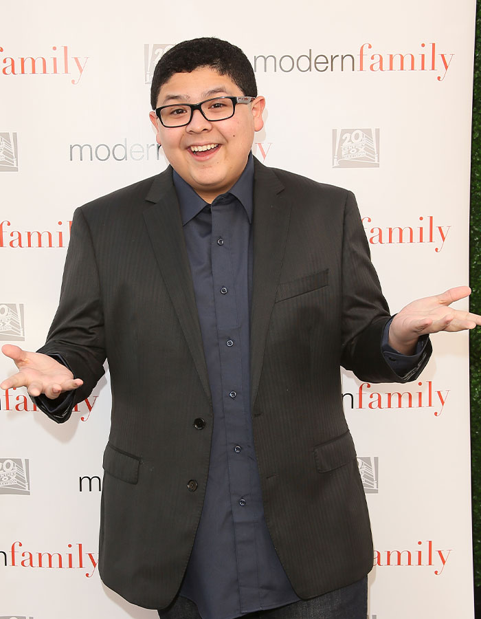 Modern Family actor in a black suit and glasses, smiling on the red carpet.
