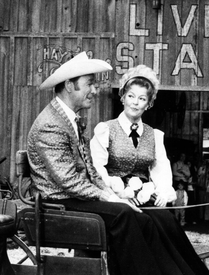 Two people in vintage western attire, sitting together, with a rustic backdrop.