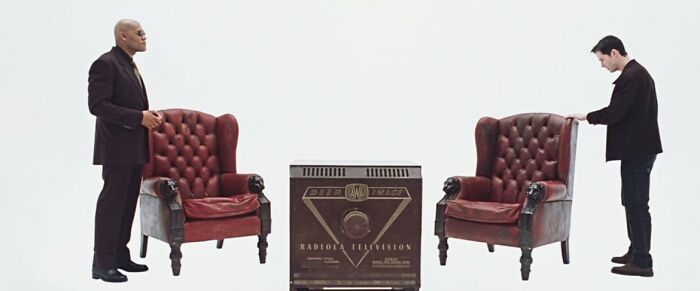 Two actors stand near vintage chairs with famous movie props on a white background.