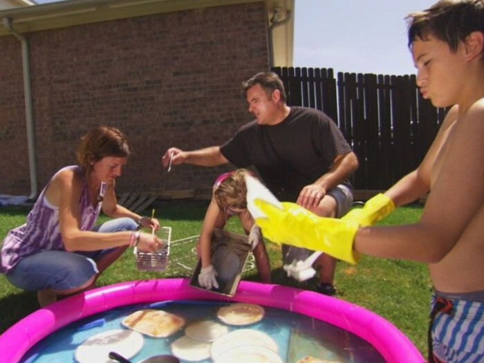 Family exercising frugality by washing dishes in a small pool, showcasing creative outdoor cleaning methods.