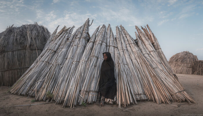 Ethiopian person standing in front of traditional reed structure, wearing a dark cloak under a blue sky.