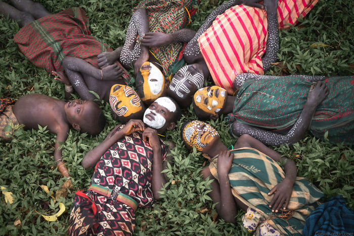 Ethiopian people in vibrant traditional outfits and painted faces, lying on green grass in a circular formation.