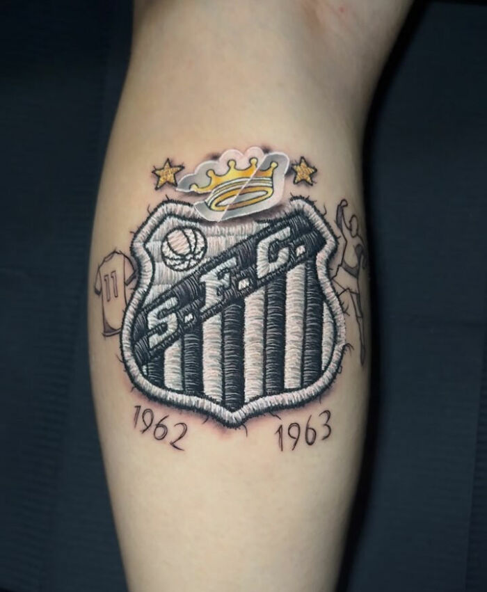 Realistic embroidery tattoo of a logo, featuring 3D thread details and surrounding elements on skin.