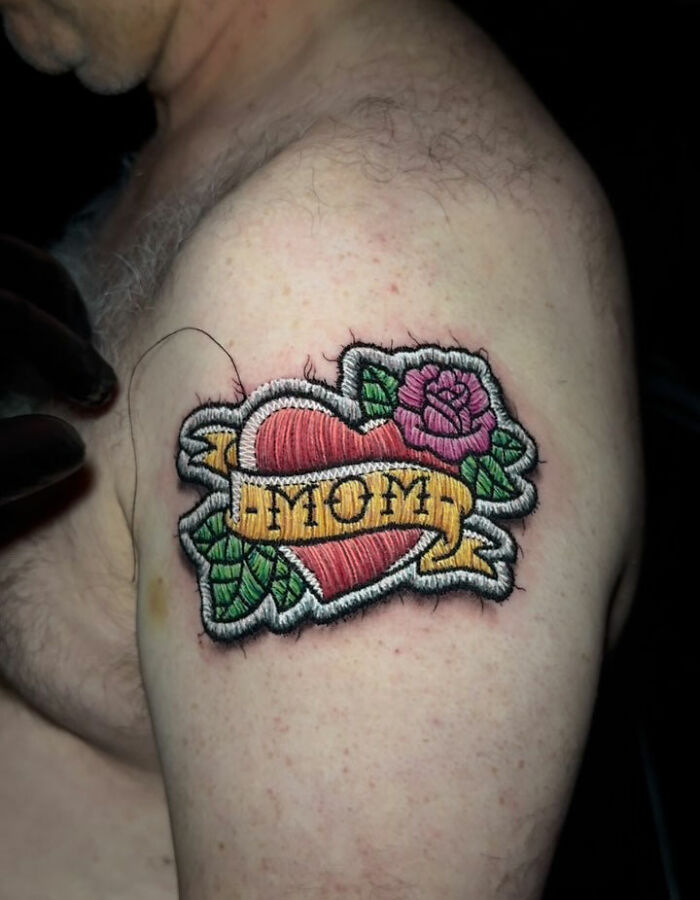 Realistic-looking embroidery tattoo of a heart and rose with "Mom" banner on upper arm.