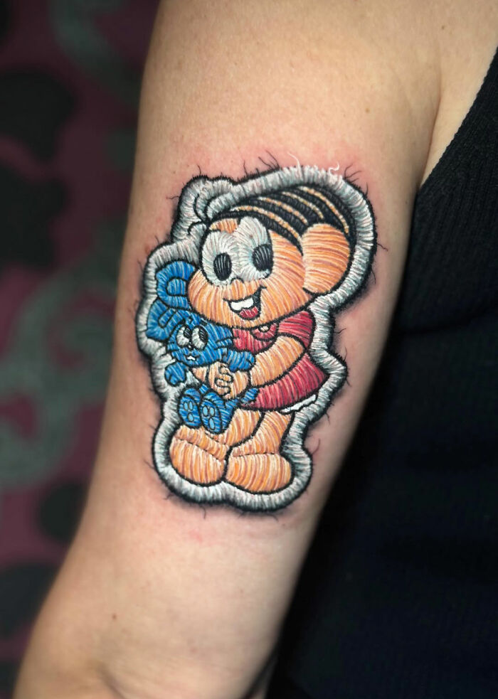 Realistic embroidery-style tattoo of a cartoon character on an upper arm.