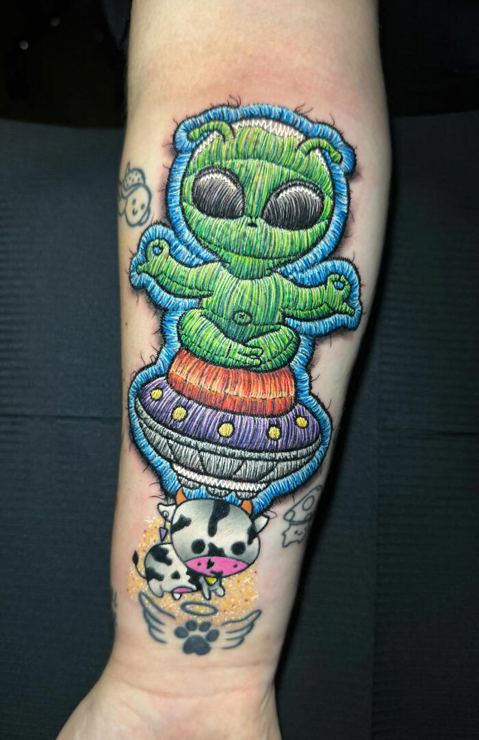 Embroidery tattoo of an alien meditating on a UFO with a cow beneath on an arm.