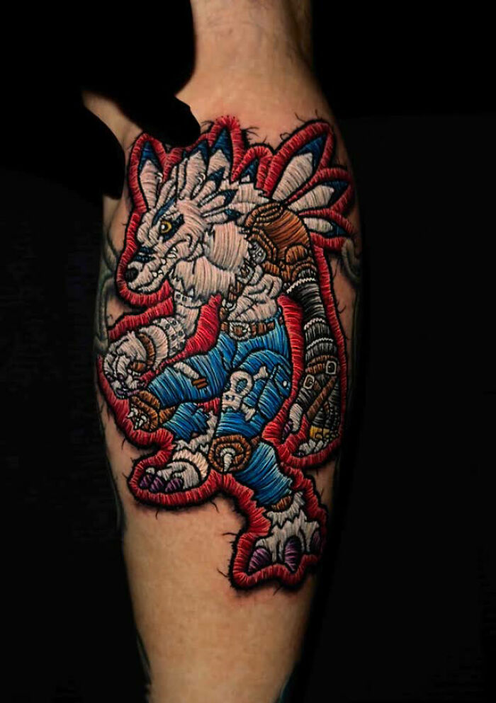 Realistic embroidery tattoo of a dinosaur on a forearm with vibrant blue, red, and white thread details.