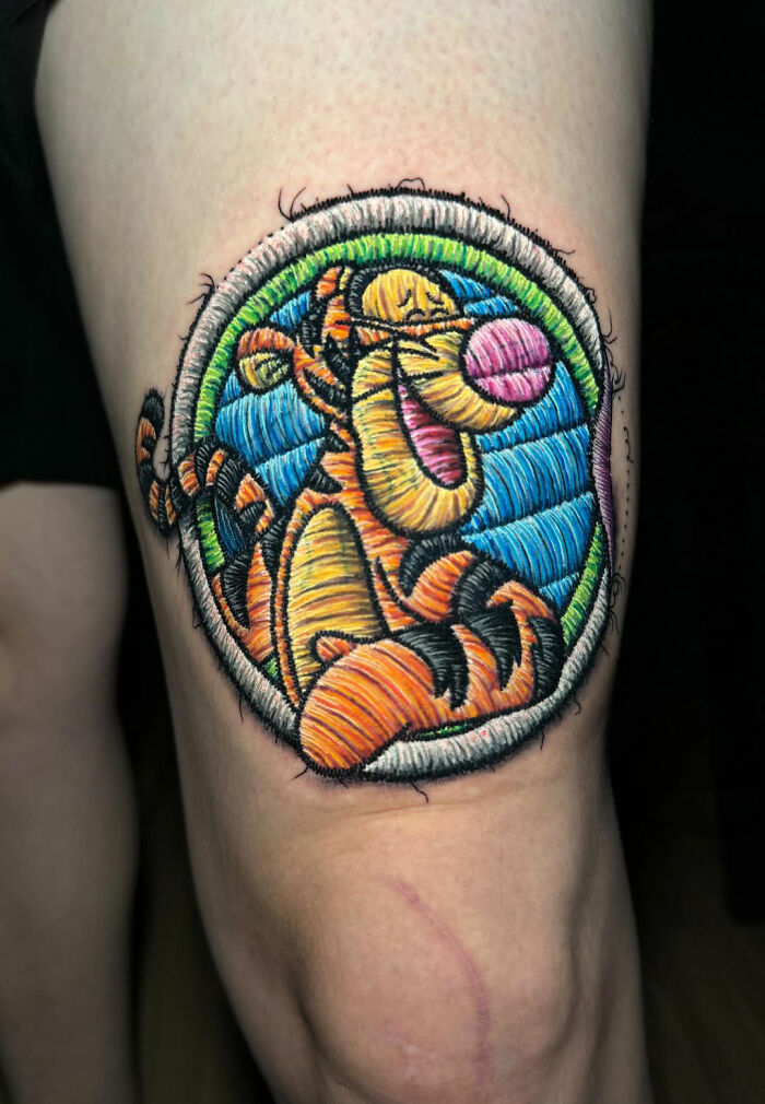Realistic embroidery tattoo of a cartoon tiger on a person's leg, showcasing vibrant colors and intricate details.