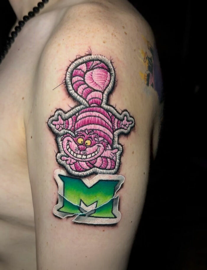 Embroidery-style tattoo of a pink cat with a green letter "M" on an arm.