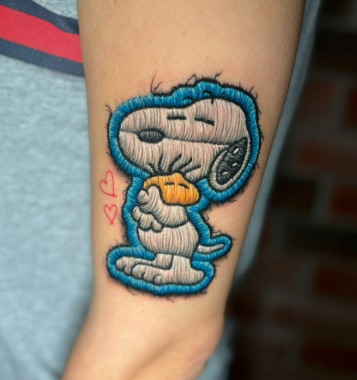 Realistic embroidery tattoo of Snoopy on arm, featuring blue outlines and small red hearts.