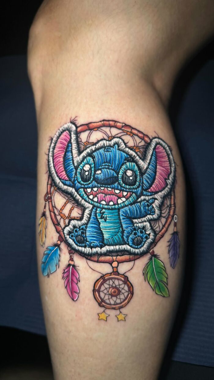 Realistic embroidery tattoo featuring a blue cartoon character with dreamcatcher elements.