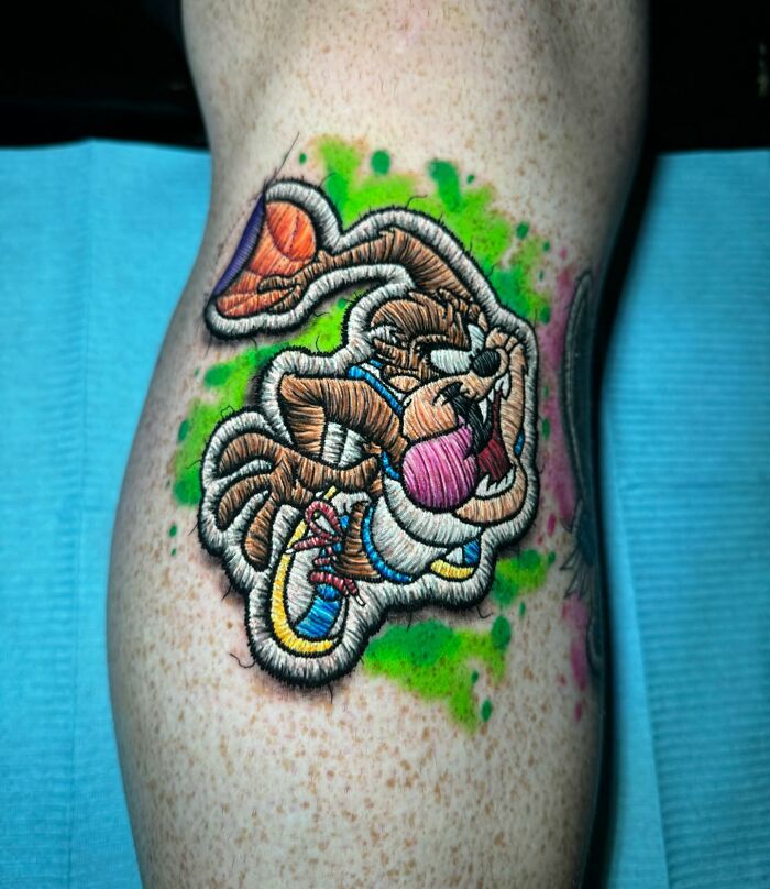 Realistic embroidery tattoo on skin, featuring a vibrant cartoon character with green background accents.