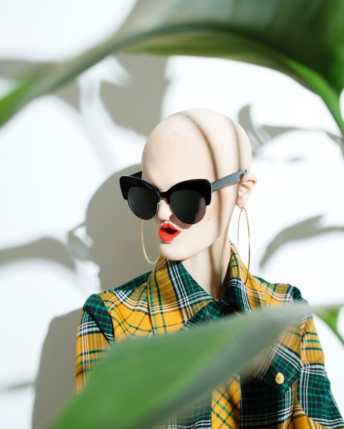 Unique model in a plaid outfit with large sunglasses and bold earrings, framed by green leaves.