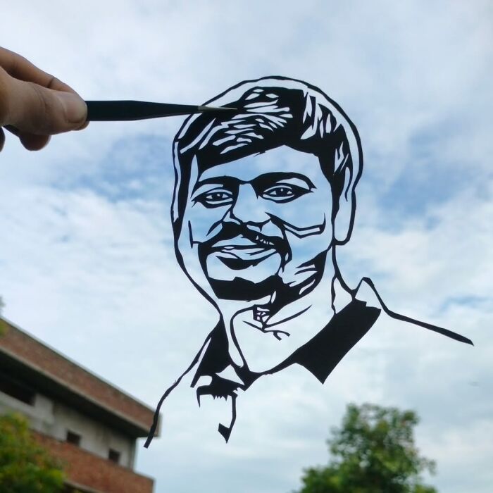 Paper-cutting artist holding a silhouetted portrait cutout against a cloudy sky.