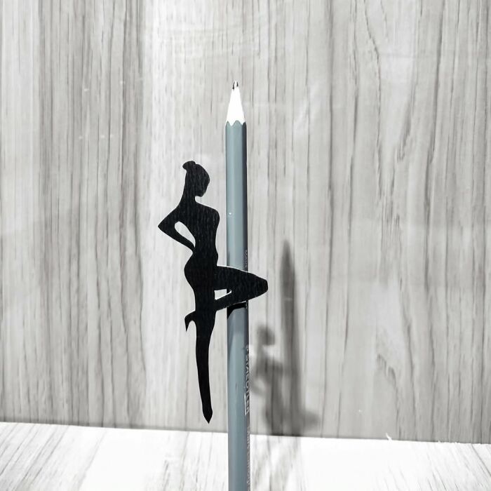 Paper-cutting art of a silhouette balancing on a pencil, showcasing intricate design against a wooden background.