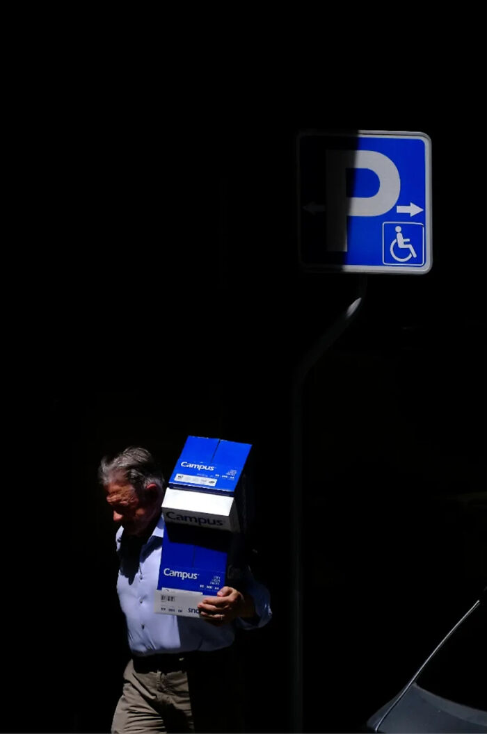 Abstract street photograph featuring a man carrying boxes under a parking sign with dramatic lighting.