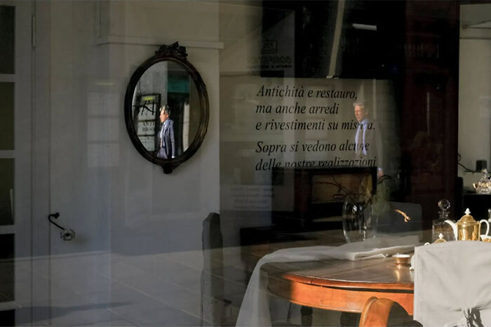 Abstract street photograph showing a reflection of a person in an ornate mirror, with text visible on the window.