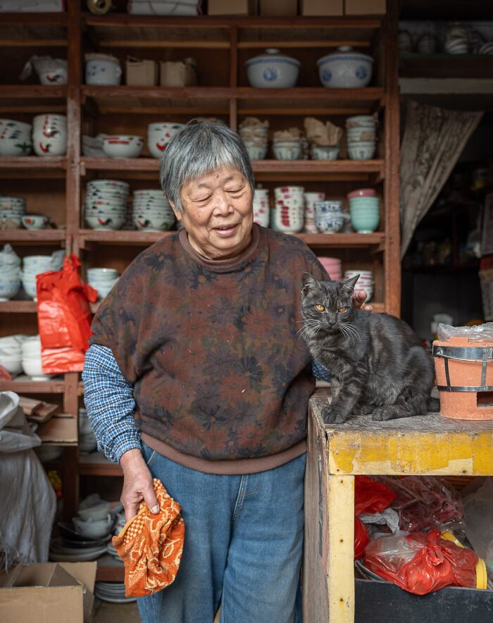Elderly woman with a cat in a cozy urban shop setting, surrounded by ceramic bowls and pots.