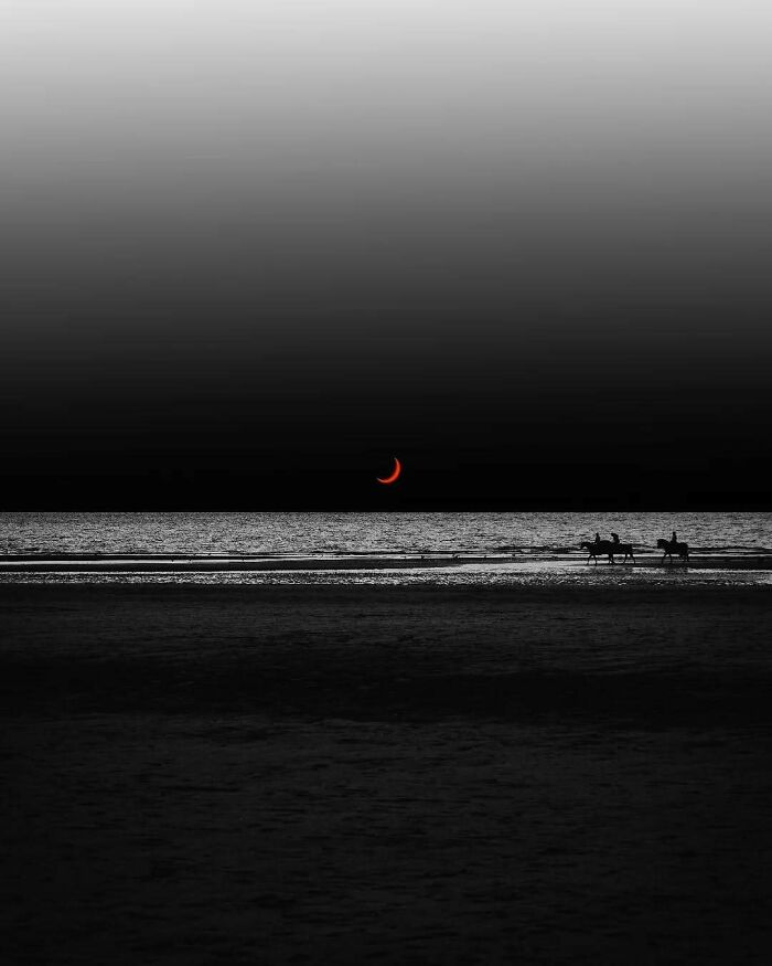 Silhouetted horse riders on a beach at night, with a surreal red crescent moon above.